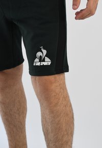 Pantalones cortos deportivos negros hechos de un material suave, con un logo blanco de "Le Coq Sportif" y un diseño de franjas laterales.