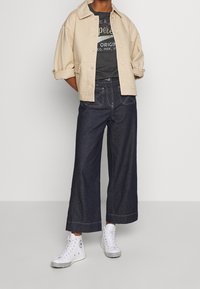 Beige quiltet jakke med krave, grå grafisk t-shirt, mørke brede bukser og hvide høj-top sneakers med sorte accenter.