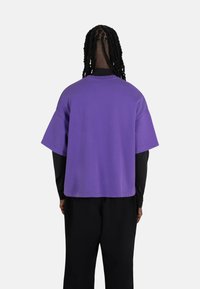 Lilla oversized t-skjorte med korte ermer, med en løs passform og minimale sømdetaljer, brukt over en svart langermet topp.