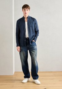Navy linen button-down shirt, white undershirt, faded blue denim jeans, white sneakers, standing on wooden floor with gray backdrop.