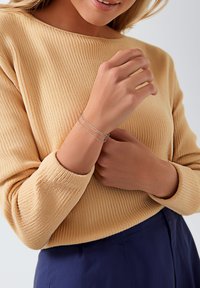 Pulsera de plata con varias capas en una muñeca, que presenta cadenas unidas. La persona que la lleva puesta lleva un suéter de punto beige, que resalta los detalles de la pulsera.