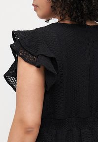 Black dress with intricate lace texture and ruffled sleeves, featuring a fitted bodice and a relaxed, flowing skirt.