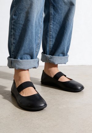 Persona con vaqueros azules remangados y bailarinas negras estilo Mary Jane con tiras elásticas, de pie sobre un suelo de hormigón claro.