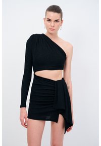 Černý top na jedno rameno s řaseným designem, který má výřez a draped sukni svázanou v pase. Materiál má jemný lesk.