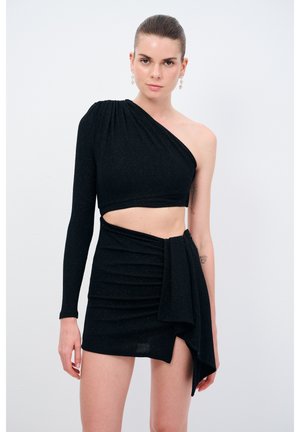 Vestido negro de un solo hombro que presenta un top corto ajustado y una minifalda fruncida con detalle de lazo lateral. La tela tiene un brillo sutil.