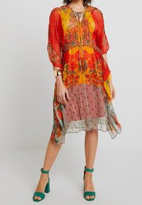 Abito trasparente con motivi floreali rossi, gialli e verdi, vita arricciata e dettaglio con cintura. Indossato con sandali con tacco verdi.