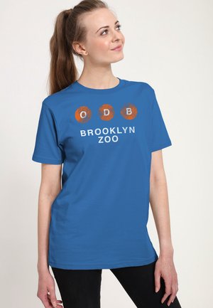 Kvinde med lys hud og langt brunt hår bærer en blå t-shirt med orange cirkler og tekst, der lyder ODB Brooklyn Zoo, kigger op og smiler let.