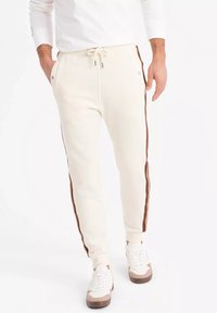 Pantalones de chándal blancos con franjas laterales marrones, con cintura ajustable con cordón, bolsillos laterales y puños acanalados. Material suave, diseño casual.