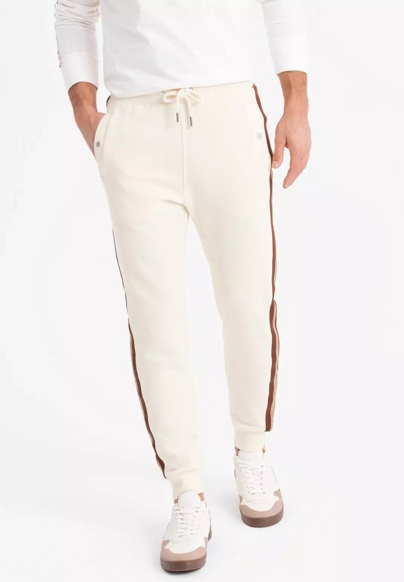 Pantalones de chándal blancos con franjas laterales marrones, con cintura ajustable con cordón, bolsillos laterales y puños acanalados. Material suave, diseño casual.