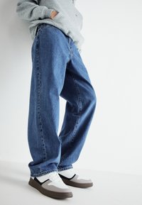 Vaqueros de mezclilla azul con un corte holgado, que cuentan con cinco bolsillos, costuras visibles y un dobladillo vuelto. Combinados con zapatillas blancas y grises.