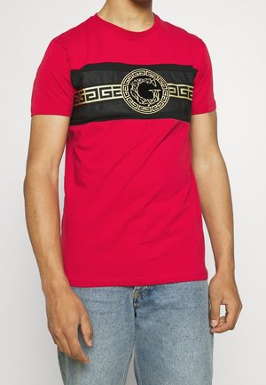 Homme portant un t-shirt rouge avec une bande de motif clé grecque noire et dorée et un emblème circulaire sur la poitrine, associé à un jean bleu clair.