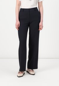Pantalons large en marine avec une texture lisse, une taille plate et des passants de ceinture, associés à des chaussures légères à enfiler avec des accents matériels.