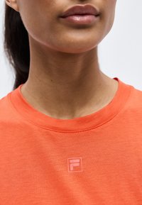 Gros plan d'une personne portant un t-shirt orange à col rond avec un petit logo blanc en forme de F sur la poitrine.