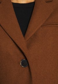 Brun uldblandet frakke med slidslapel, enkelt knaplukning og blød tekstur, med en sort ribbet top under.