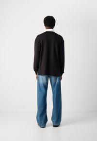 Persona con capelli ricci corti che indossa una maglietta nera a maniche lunghe sopra una camicia con colletto bianco e jeans blu larghi, in piedi davanti a uno sfondo neutro.