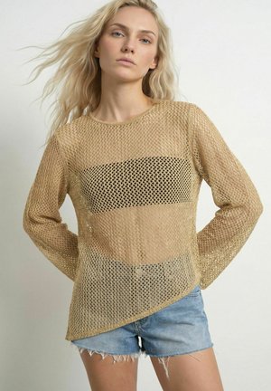 Kvinde med blondt hår bærer en tan åbenhullet langærmet top over en sort bandeau og lyseblå denimshorts.