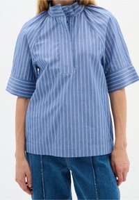 Blårandig skjorta med hög krage, korta ärmar och knäppning fram. Har vertikala vita ränder och ett bröstfack fram.