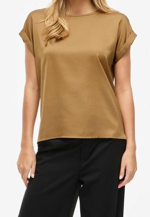 Vrouw draagt een camelkleurige blouse met korte mouwen en opgerolde manchetten en een zwarte broek tegen een effen achtergrond.