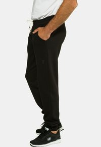 Zwarte joggingbroek met een tailleband en trekkoord, zijzakken en een reliëflogo op het been, gecombineerd met zwarte sportschoenen.