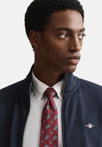 Veste zippée bleu marine avec une chemise blanche en dessous et une cravate bordeaux à motif paisley. La cravate présente des motifs bleus et jaunes ainsi qu'un logo GANT.