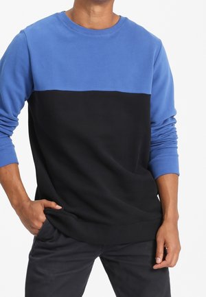 Mann trägt langärmeligen Sweatshirt mit blauem Oberteil und schwarzem Unterteil, Hand in der Tasche, steht vor weißem Hintergrund.