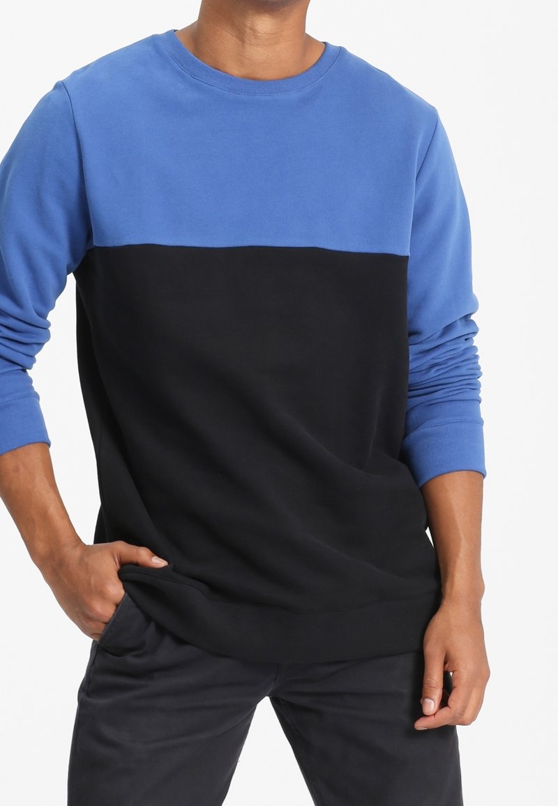 Homme portant un sweat-shirt à manches longues avec le haut bleu et le bas noir, main dans la poche, debout devant un fond blanc.