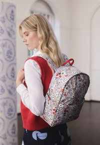 Szőke nő vörös ujjatlan pulóverben fehér hátizsákot visel, amelyet kártyalapok és virágminta díszítenek, beltéren.