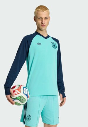 Jonge man in een voetbaltenue in groenblauw en marineblauw, met een meerkleurige voetbal in de handen, staand tegen een effen lichte achtergrond.