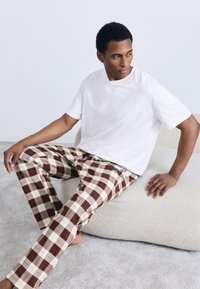 Weißes T-Shirt mit Rundhalsausschnitt, kombiniert mit braun-cremefarben karierter Hose, mit lockerem Schnitt und glattem Stoff, sitzend auf einer weichen Oberfläche.