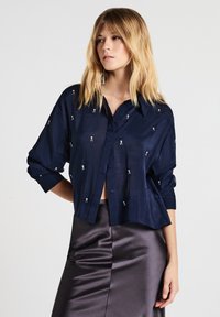 Una donna bionda che indossa una camicia blu navy con bottoni e piccoli disegni ricamati bianchi, insieme a una gonna di raso grigio scuro lucido.