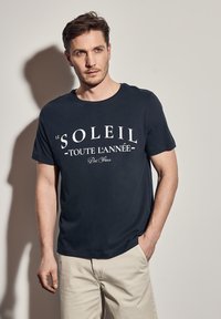 Tmavě modré tričko s krátkým rukávem a kulatým výstřihem. Obsahuje bílý text s nápisem "Le Soleil Toute L'Année Port Fleux." Hladká textura.