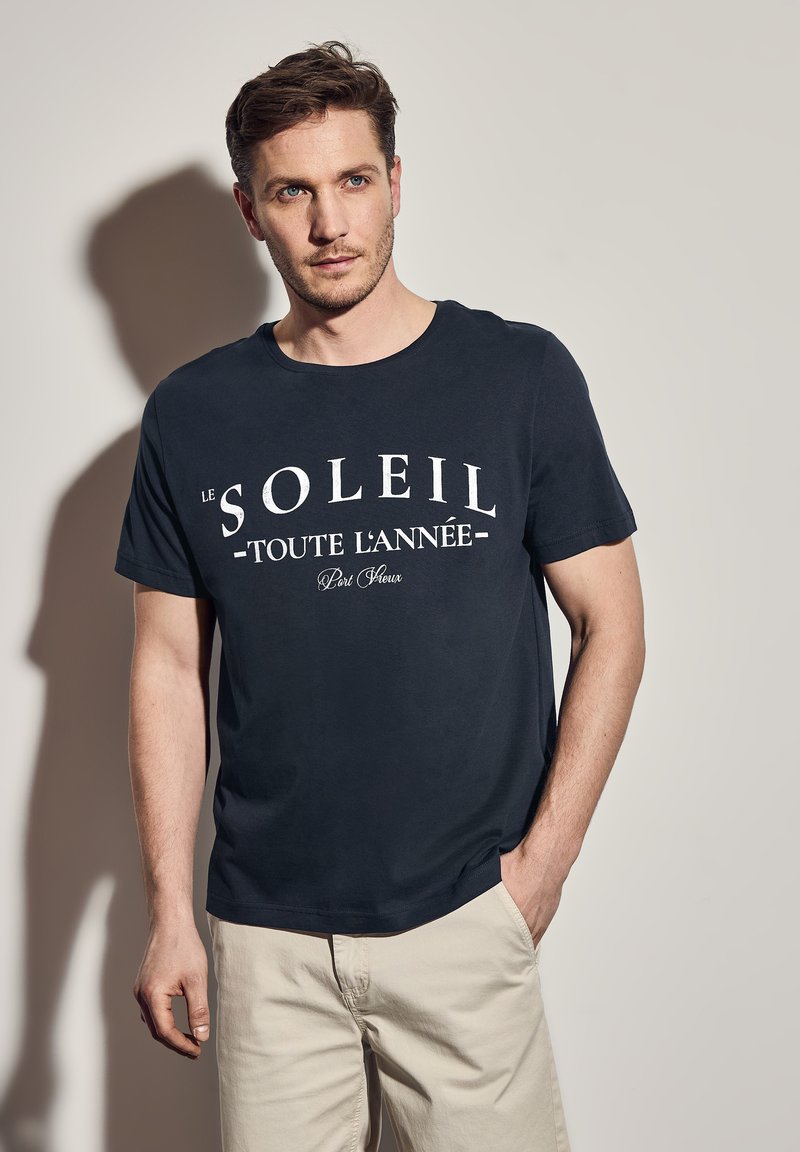 Tmavě modré tričko s krátkým rukávem a kulatým výstřihem. Obsahuje bílý text s nápisem "Le Soleil Toute L'Année Port Fleux." Hladká textura.