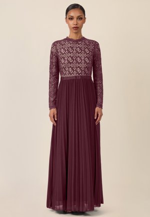 Frau mit glattem Haar trägt ein bodenlanges, kastanienbraunes Kleid mit einem Oberteil aus Spitzenmuster und einem langen, plissierten Rock, steht vor einem beigen Hintergrund.