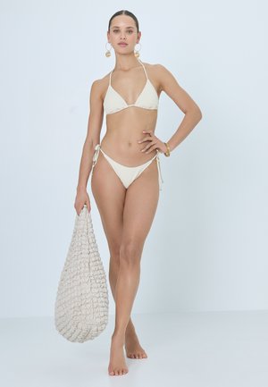 Frau trägt einen elfenbeinfarbenen, strukturierten String-Bikini, große Creolen und Armbänder, hält eine große elfenbeinfarbene geflochtene Tasche, steht barfuß.