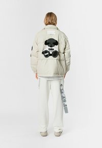 Chaqueta acolchada beige con un gran bordado de un oso negro en la espalda, que presenta una textura suave y detalles de cremallera. Combinada con pantalones blancos.