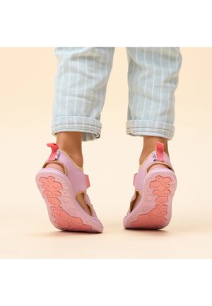 Kind draagt lichtblauwe opgerolde broek en roze sandalen met getextureerde zolen, staand op een beige achtergrond.
