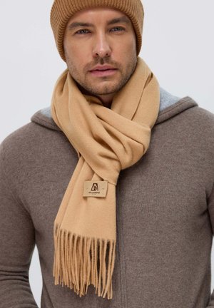 Sciarpa di lana beige con dettaglio a frange, dotata di un'etichetta con logo. Indossata sopra un maglione marrone con cappuccio, abbinata a un berretto coordinato.