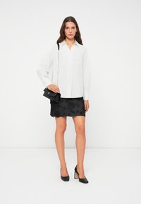 Camicia bianca a bottoni, mini gonna nera texturizzata con motivo floreale, borsa nera con tracolla a catena e scarpe con tacco nere.