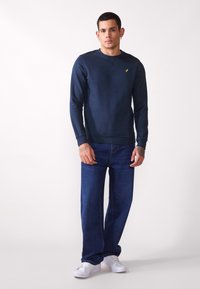 Navy blue long-sleeve sweatshirt with round neckline, textured fabric, and a small yellow logo on the chest, paired with blue jeans and white sneakers.