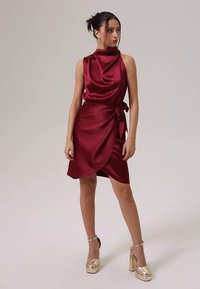 Bordinės spalvos satino suknelė su aukštu kaklu, klostuotu dizainu, surištu juosmeniu ir apsiaustine siluetu. Derinama su auksinės spalvos platformos kulnais.