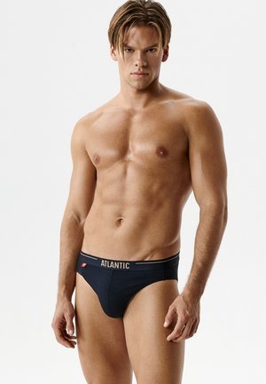 Młody męski model ubrany w czarne markowe bokserki z napisem "ATLANTIC" na pasie, stojący na tle jednolitej białej ściany, zwrócony przodem.