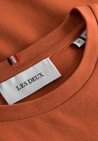 Rustfarget t-skjorte med rund hals, med en hvit etikett som leser "LES DEUX" og små røde, hvite og blå detaljer nær kragen.
