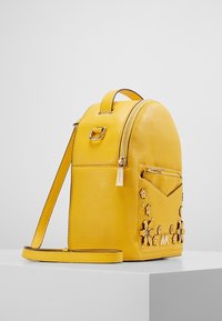 Mochila de cuero amarilla con parte superior redondeada, bolsillo frontal, adornos florales y correa ajustable. Presenta acentos de hardware dorado.