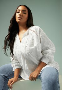 Blusa bianca ricamata con scollatura a V e dettagli con frange, abbinata a jeans in denim azzurro. Il tessuto appare testurizzato con motivi floreali.