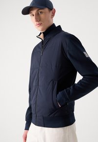 Navy zip-up jacket featuring a padded front, knit sleeves, ribbed cuffs, and a small logo patch on the left arm.