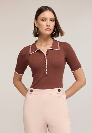 Vrouw met steil bruin haar draagt een bruine geribbelde top met korte mouwen en witte rand, en beige broek met hoge taille en zwarte knoopdetails.