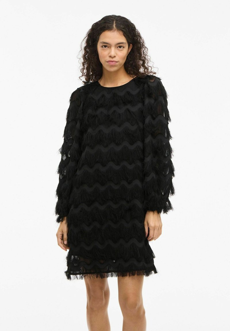 Robe noire avec des détails de frange ondulée, manches longues et encolure ronde. Le tissu transparent à l'ourlet et aux poignets ajoute de la texture et de la profondeur.