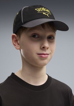 Chłopiec w czapce z logo Pokémon i Puma oraz czarnej koszulce, patrzący lekko na bok z neutralnym wyrazem twarzy.