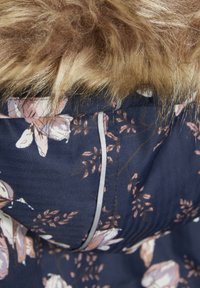 Tissu bleu marine à motif floral avec bordure en fausse fourrure beige. Présente une fermeture éclair visible et des coutures, mettant en valeur les détails et la texture du vêtement.