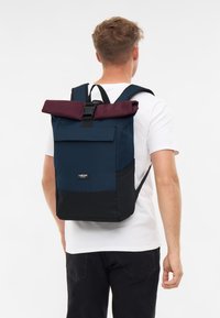 Tengerészek hátizsákja bordó, felgombolható felső résszel és fekete aljjal. Állítható pántokkal és elülső zsebbel rendelkezik, tartós anyagból készült.